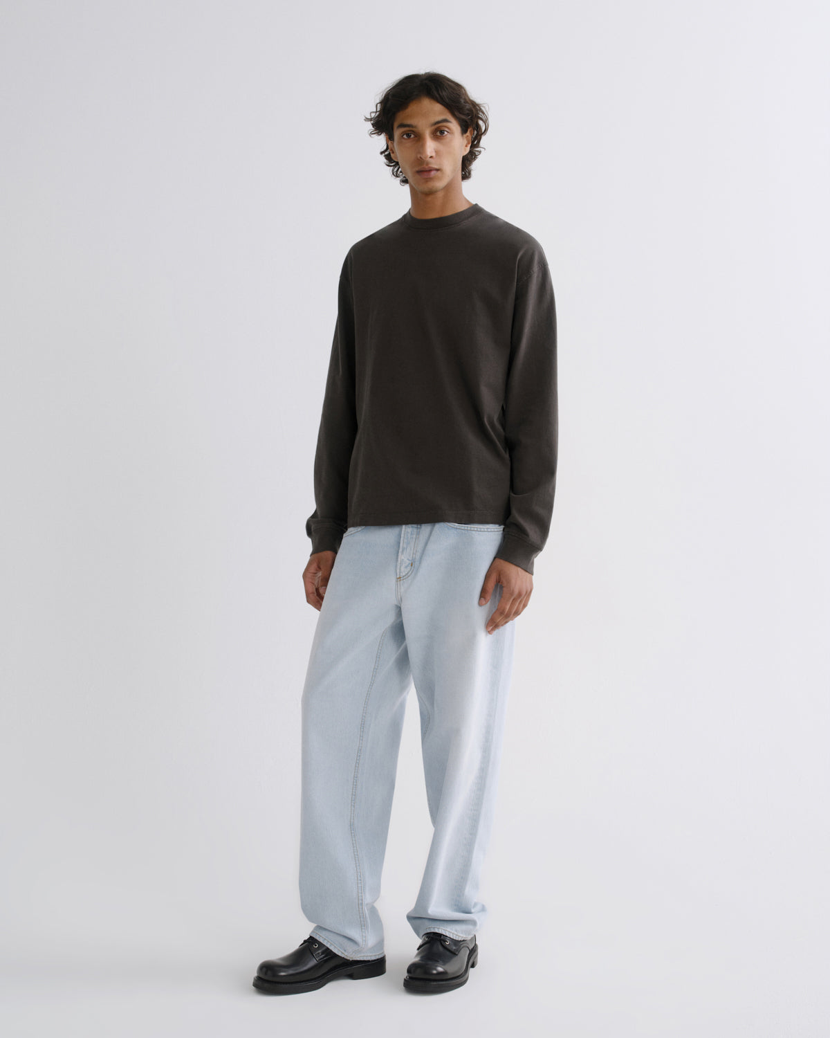 A model stands against a light gray studio background wearing a dark brown long-sleeve crewneck sweatshirt, light-wash wide-leg jeans, and black leather shoes.