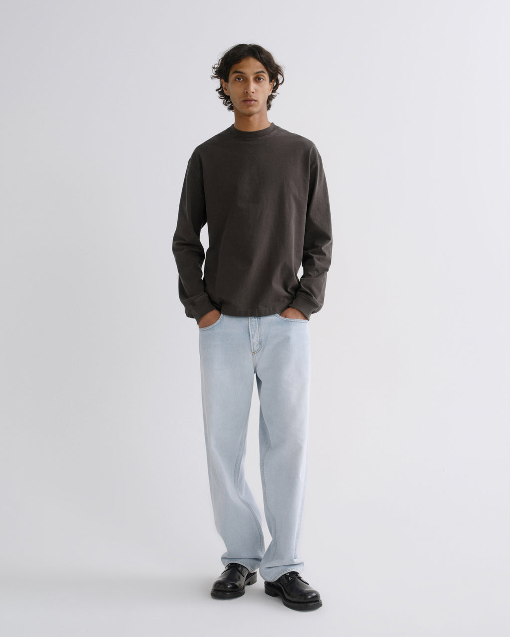 A man stands against a light gray studio background wearing a dark charcoal long-sleeve crewneck shirt, light blue jeans, and black leather shoes.