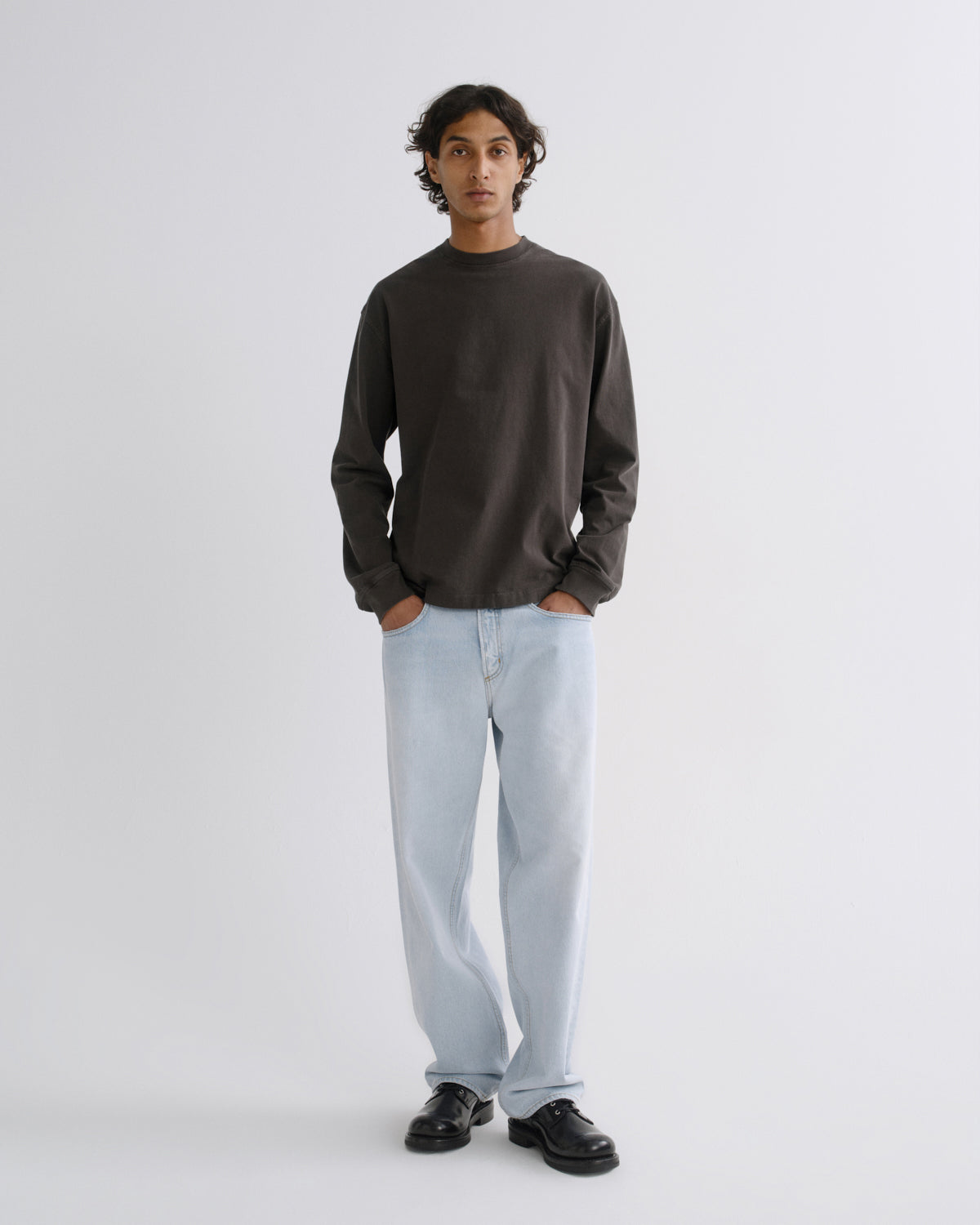 A man stands against a light gray studio background wearing a dark charcoal long-sleeve crewneck shirt, light blue jeans, and black leather shoes.