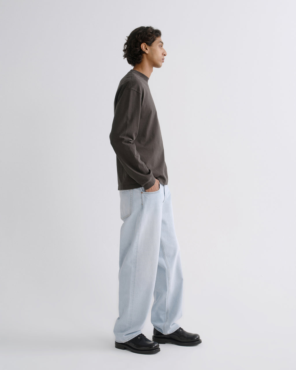 A man in profile wearing a dark charcoal long-sleeve crewneck sweatshirt, light-wash wide-leg jeans, and black leather shoes, standing against a plain light gray studio background.