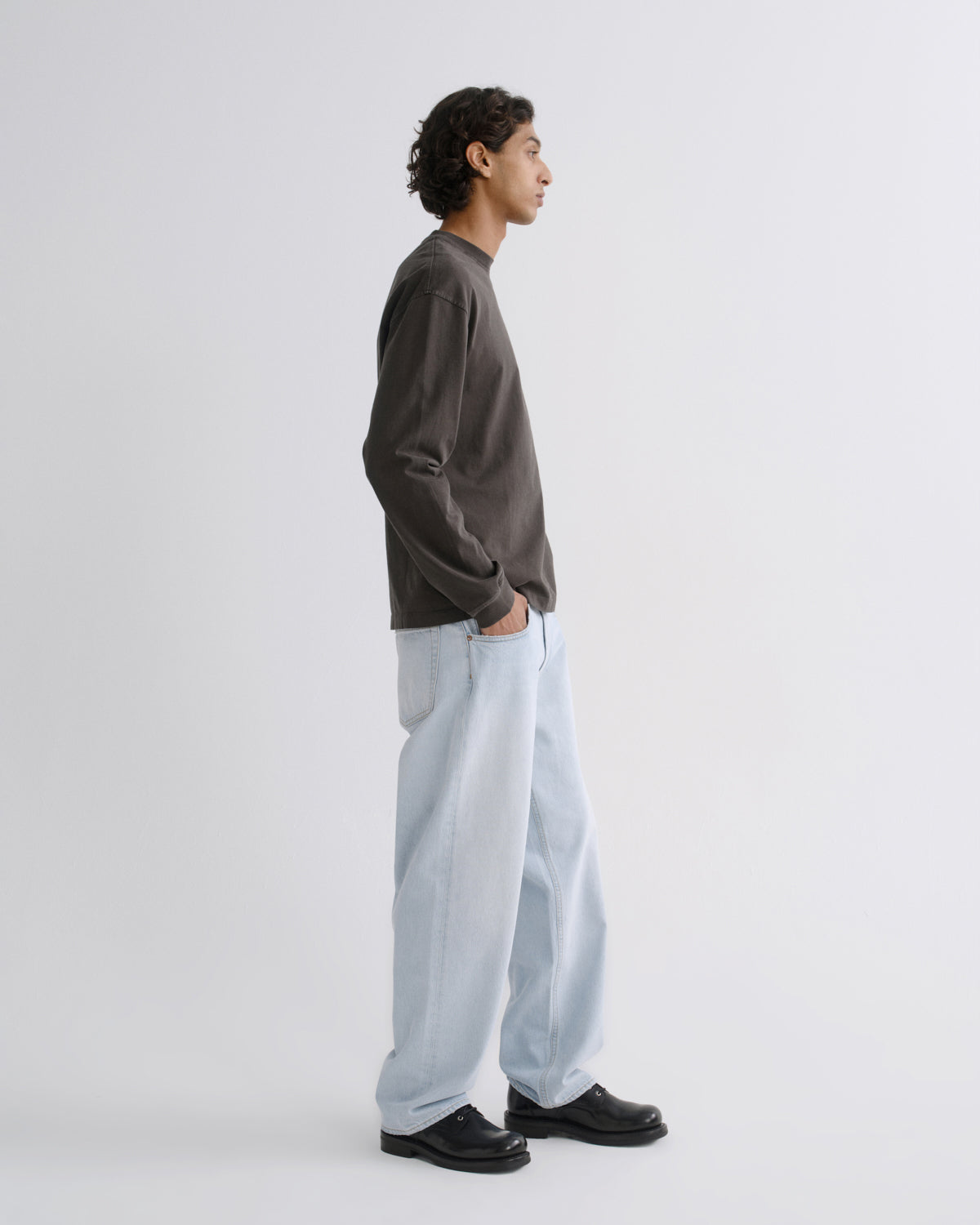 A man in profile wearing a dark charcoal long-sleeve crewneck sweatshirt, light-wash wide-leg jeans, and black leather shoes, standing against a plain light gray studio background.