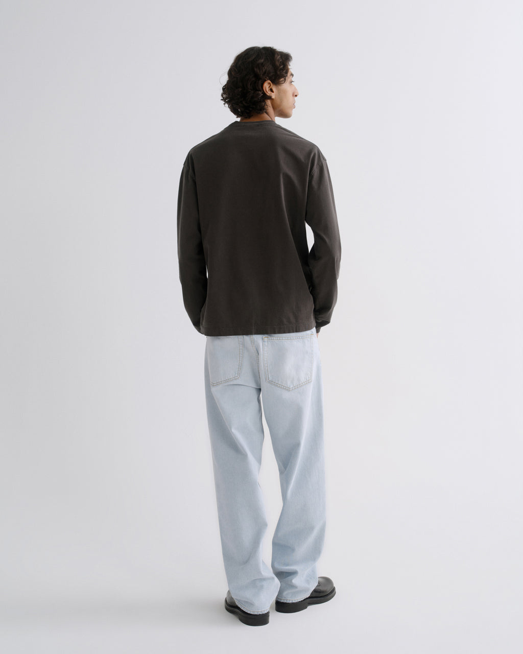 A man is shown from the back wearing a dark brown long-sleeve jacket, light blue jeans, and black shoes, standing against a plain light gray studio background.