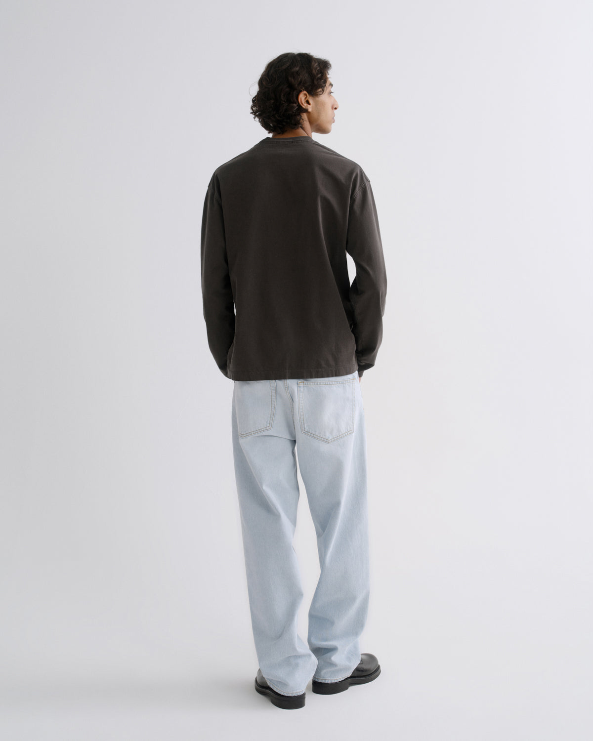 A man is shown from the back wearing a dark brown long-sleeve jacket, light blue jeans, and black shoes, standing against a plain light gray studio background.