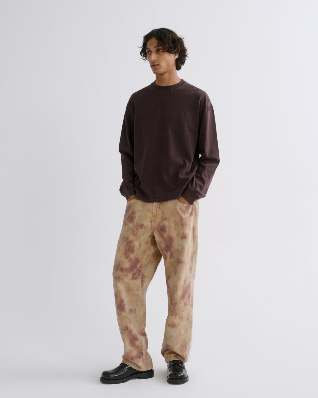 A man modeling a dark brown long-sleeve crewneck shirt and beige tie-dye pants with brown mottled patterns, paired with black leather shoes, against a plain light gray studio background.