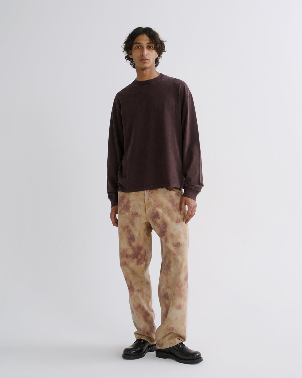 A man stands against a light gray studio background wearing a dark brown long-sleeve crewneck sweatshirt, beige pants with a brown tie-dye pattern, and black shoes.