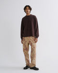 A man stands against a light gray studio background wearing a dark brown long-sleeve crewneck sweatshirt, beige pants with a brown tie-dye pattern, and black shoes.