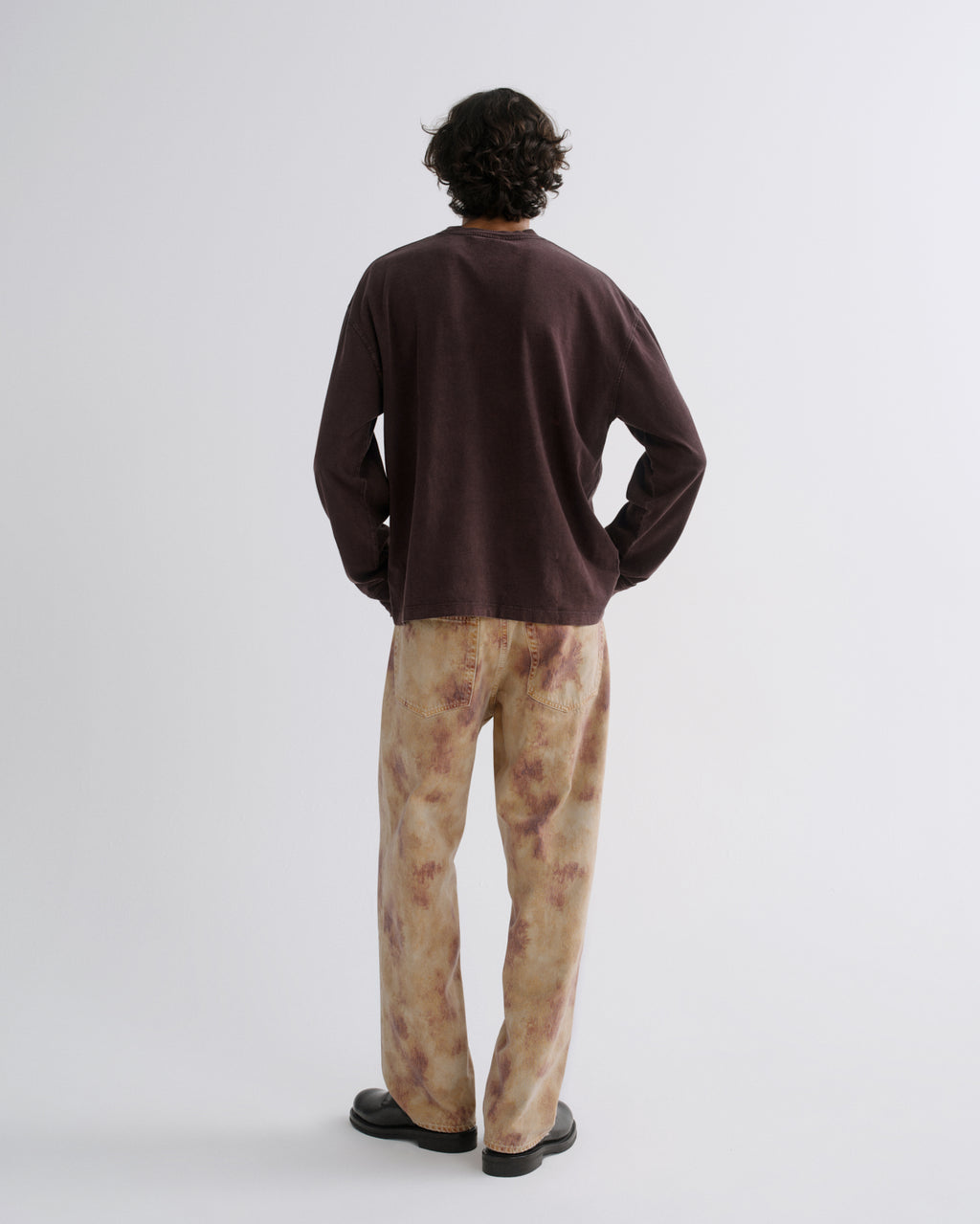 A man is shown from behind wearing a dark brown long-sleeve crewneck shirt and beige tie-dye pants, standing against a plain light gray studio background.