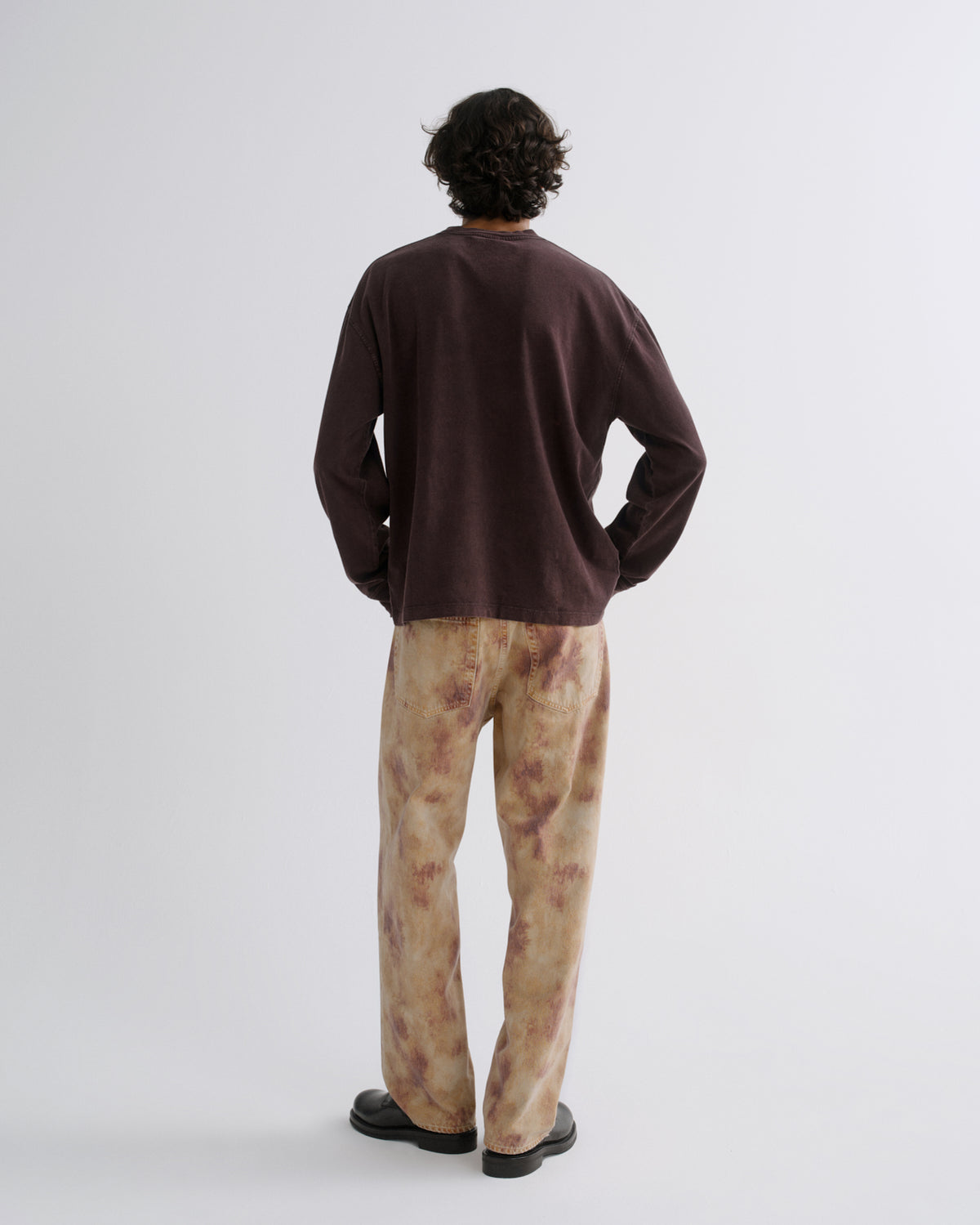 A man is shown from behind wearing a dark brown long-sleeve crewneck shirt and beige tie-dye pants, standing against a plain light gray studio background.