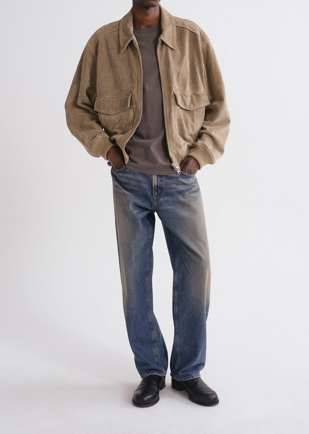 Person wearing a beige jacket, brown shirt, blue jeans, and black shoes on a grey background