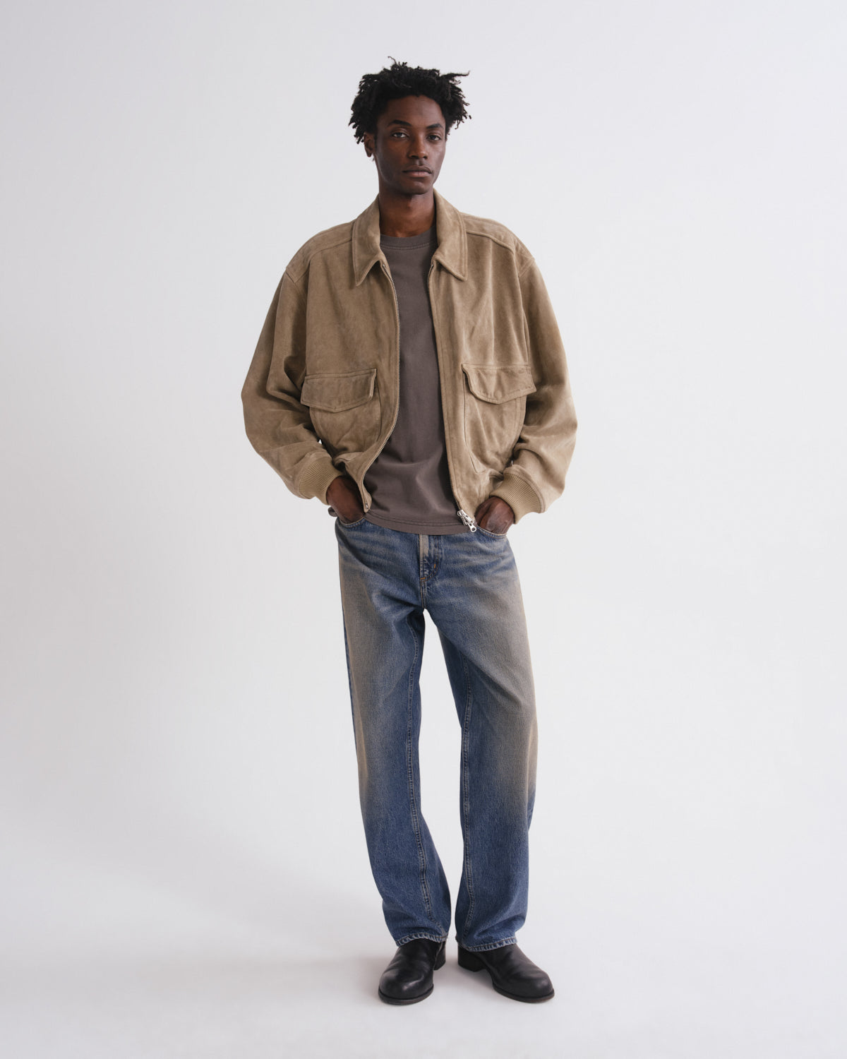 A man modeling a light brown corduroy utility jacket over a dark gray t-shirt, blue jeans, and black shoes, standing against a plain white studio background.