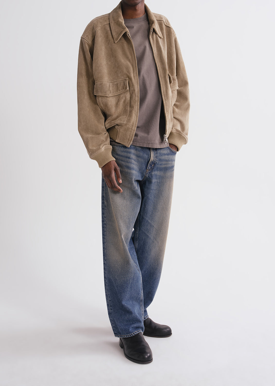 Person wearing a beige jacket, gray shirt, blue jeans, and black shoes on a grey background