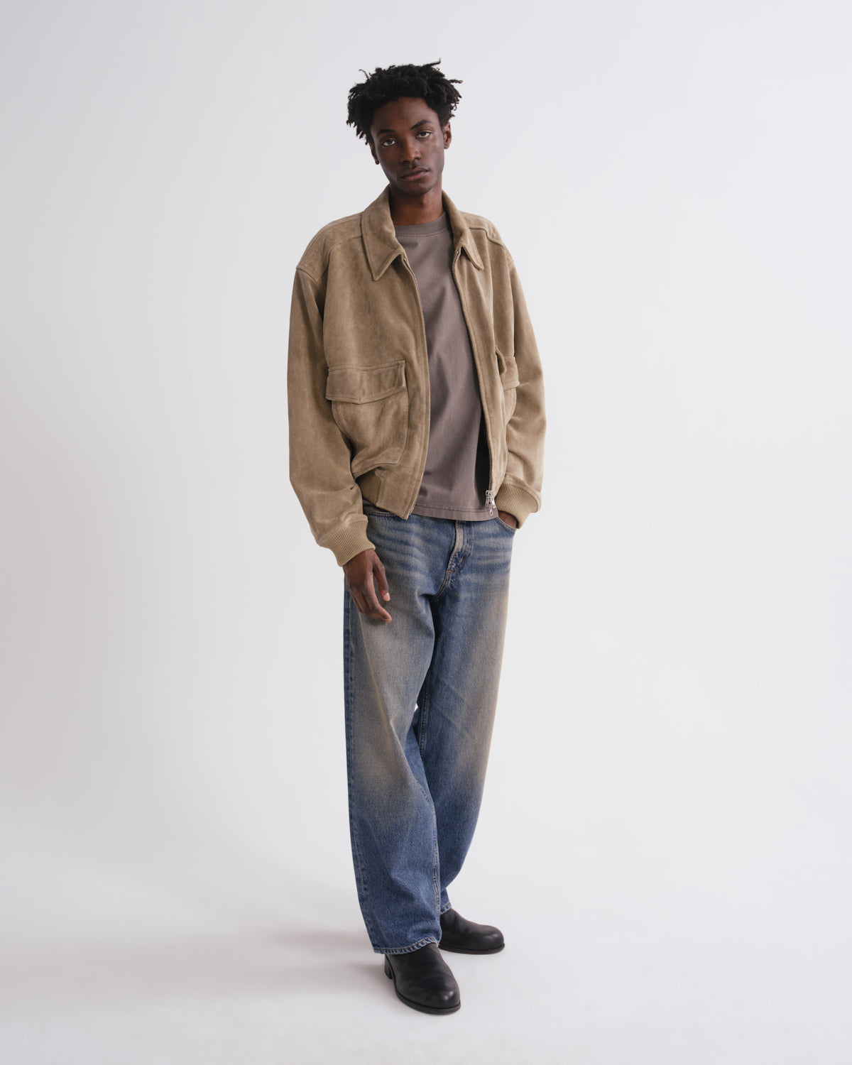 A man modeling a tan suede utility jacket over a taupe t-shirt, blue jeans, and black shoes, standing against a plain white studio background.