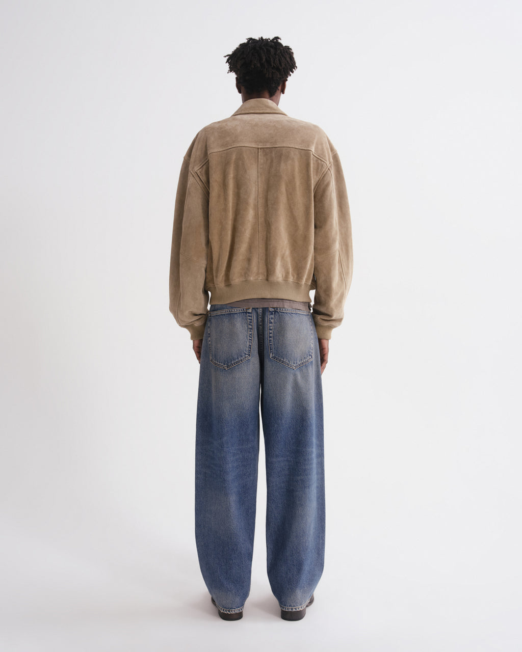 A model viewed from behind wearing a tan suede jacket and wide-leg blue jeans against a plain white studio background.