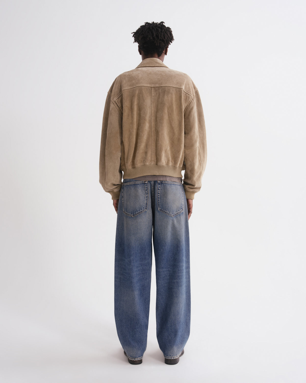 A model viewed from behind wearing a tan suede jacket and wide-leg blue jeans against a plain white studio background.