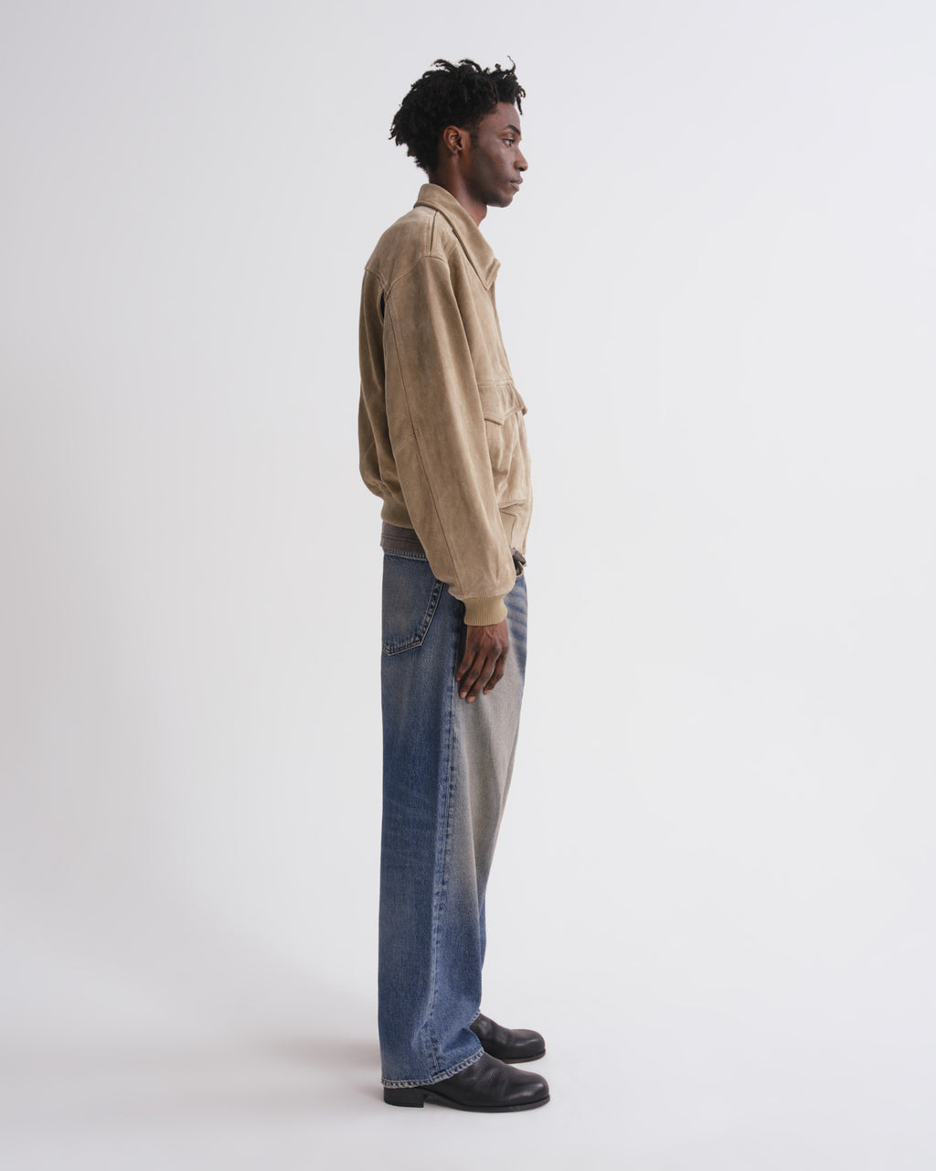 A man stands in profile wearing a beige suede jacket, wide-leg blue jeans with a light gray side panel, and black shoes against a plain white studio background.