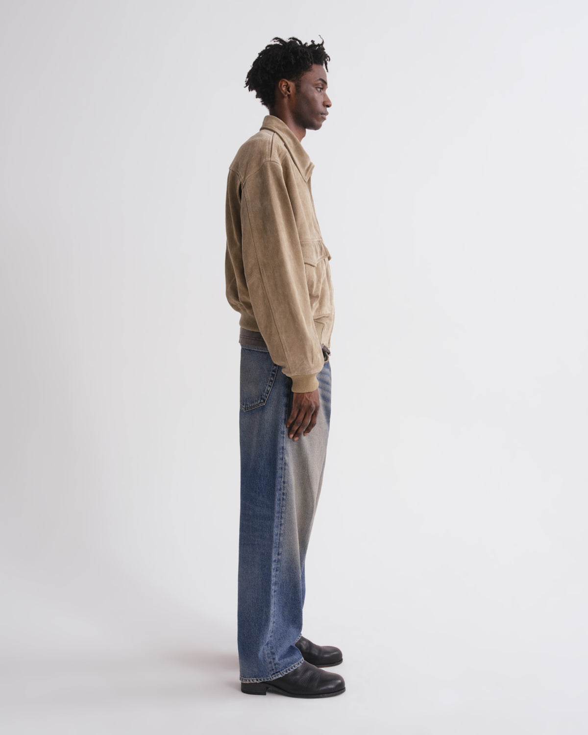 A man stands in profile wearing a beige suede jacket, wide-leg blue jeans with a light gray side panel, and black shoes against a plain white studio background.