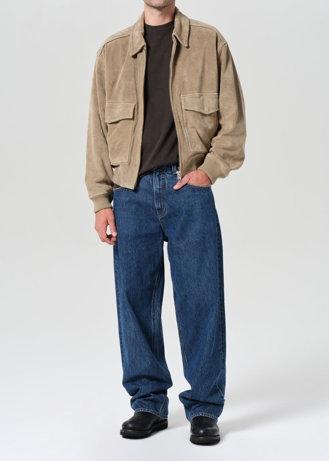 Person wearing a beige jacket, black shirt, blue jeans, and black shoes on a white background