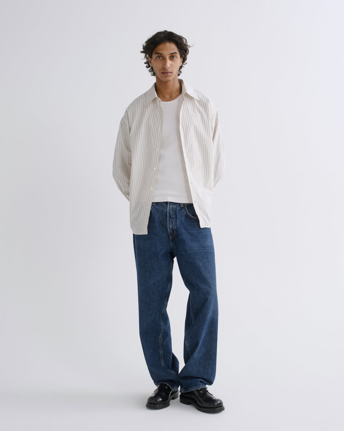 A man stands against a light gray studio background wearing a white textured button-up shirt over a white t-shirt, blue jeans, and black shoes.