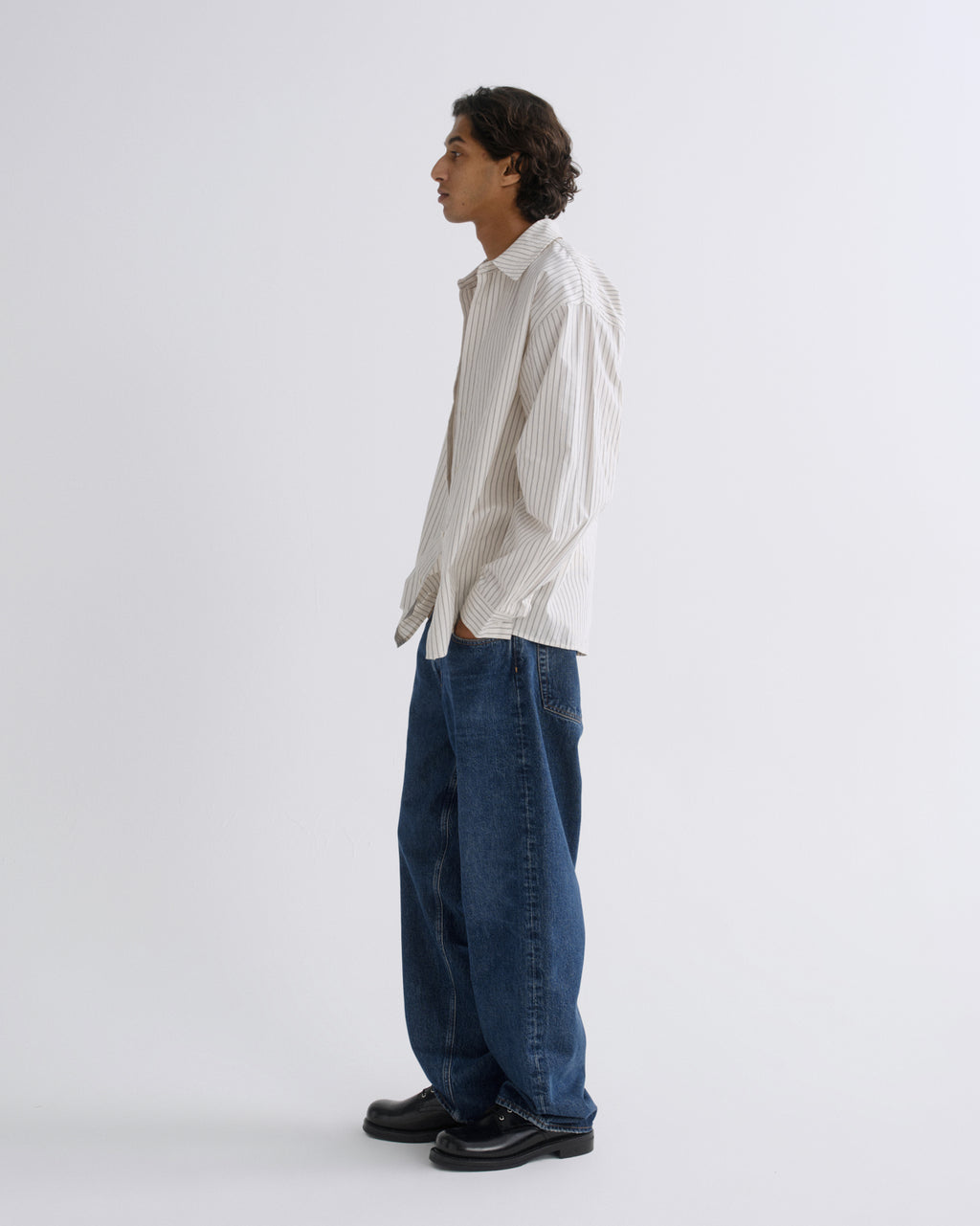 A man stands in profile wearing a cream and dark vertical-striped button-down shirt, loose-fit dark blue jeans, and black leather shoes against a plain light gray studio background.
