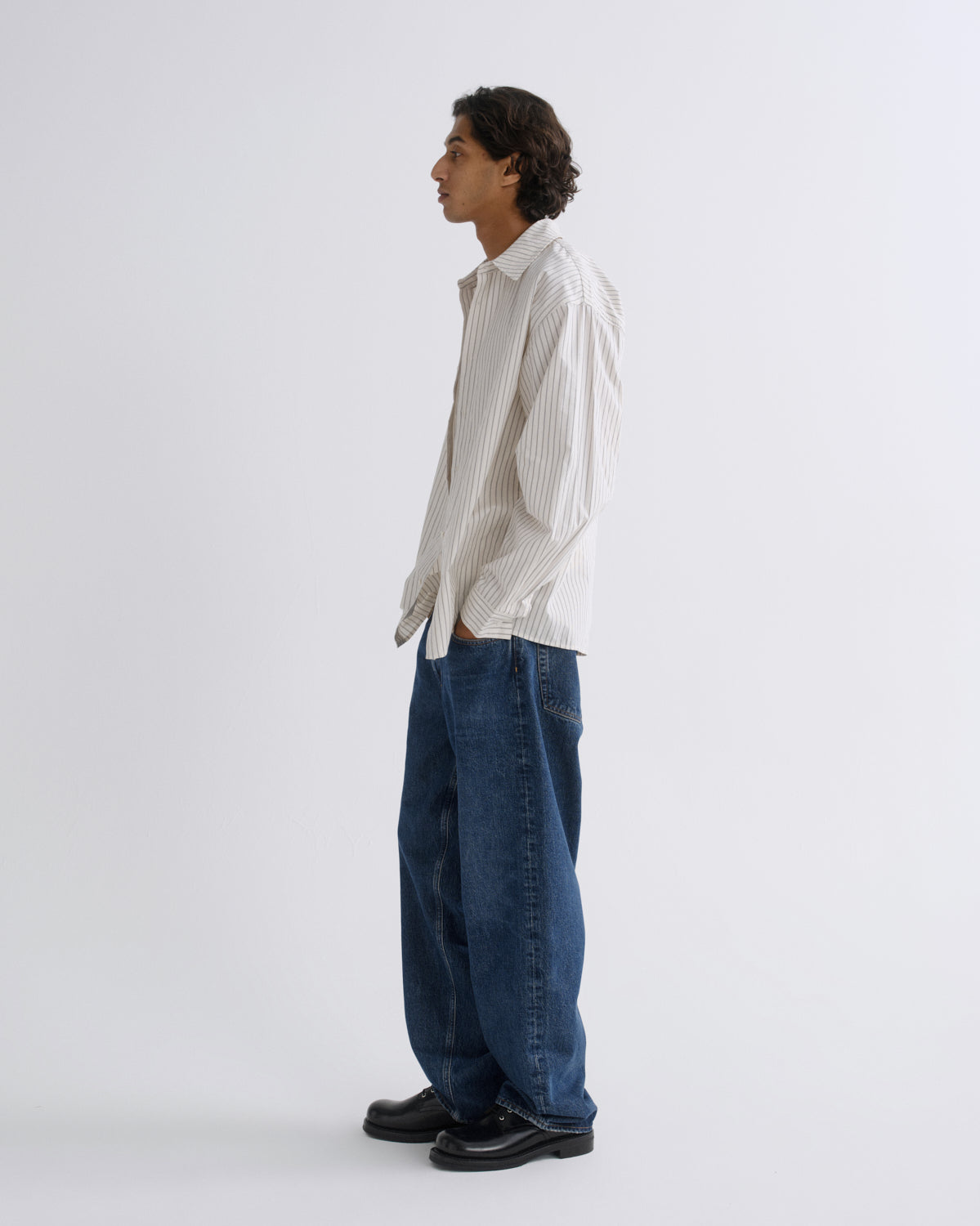 A man stands in profile wearing a cream and dark vertical-striped button-down shirt, loose-fit dark blue jeans, and black leather shoes against a plain light gray studio background.