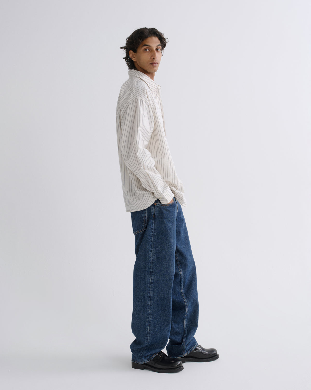 A model wearing a cream-colored vertically striped button-down shirt, loose-fit blue jeans, and black leather loafers, standing in profile against a light gray studio background.