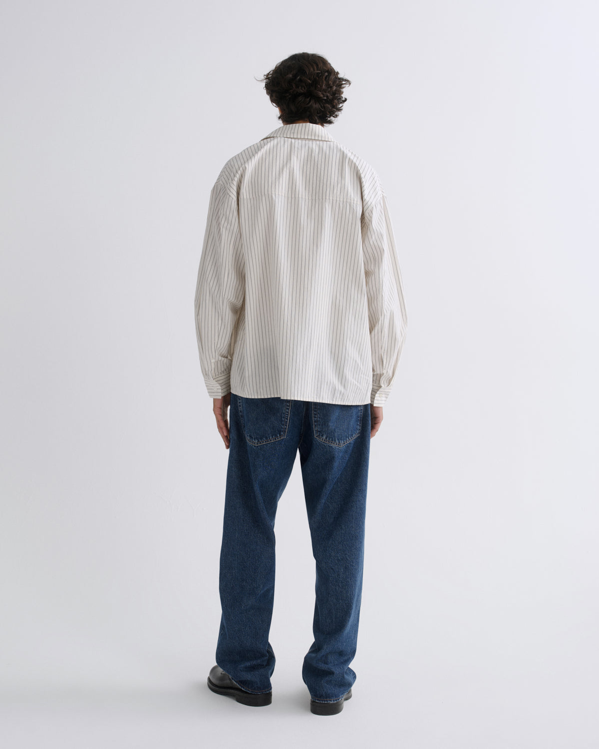 A man viewed from behind wearing a cream-colored, long-sleeve button-up shirt with thin vertical dark pinstripes, paired with blue jeans and black shoes, against a plain light gray studio background.