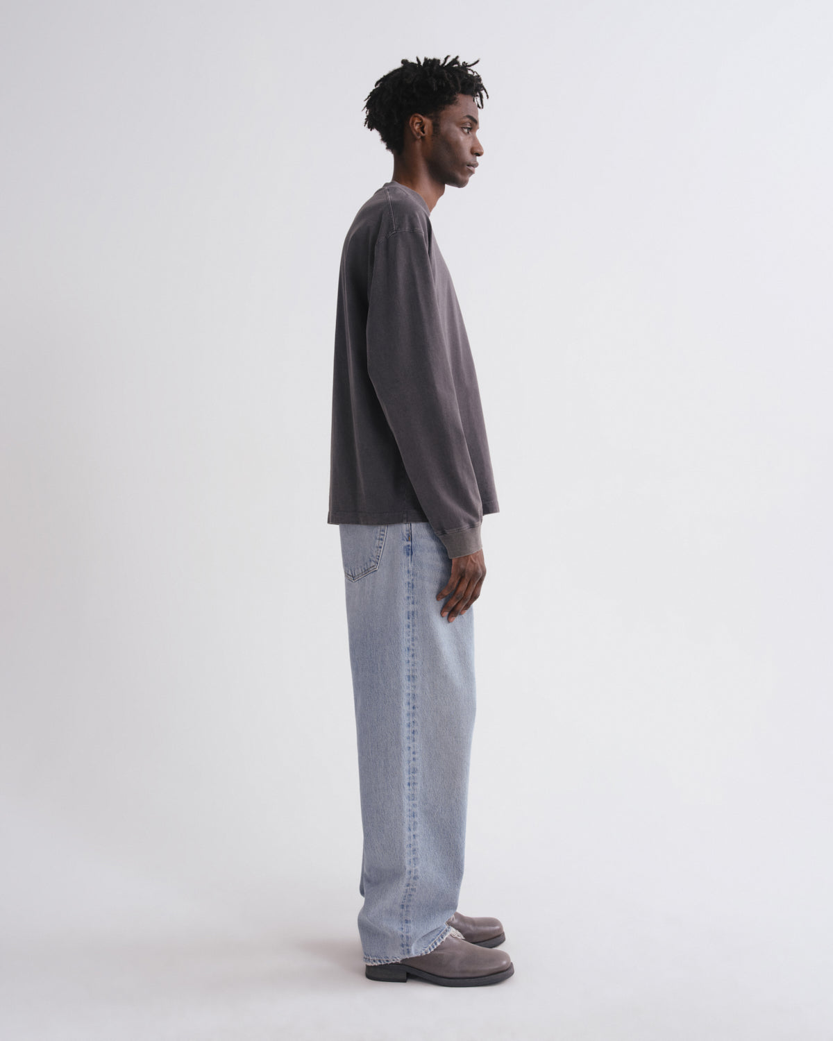 A man in profile wearing a dark gray oversized long-sleeve shirt, light-wash wide-leg jeans, and gray slip-on shoes, standing against a plain light gray studio background.