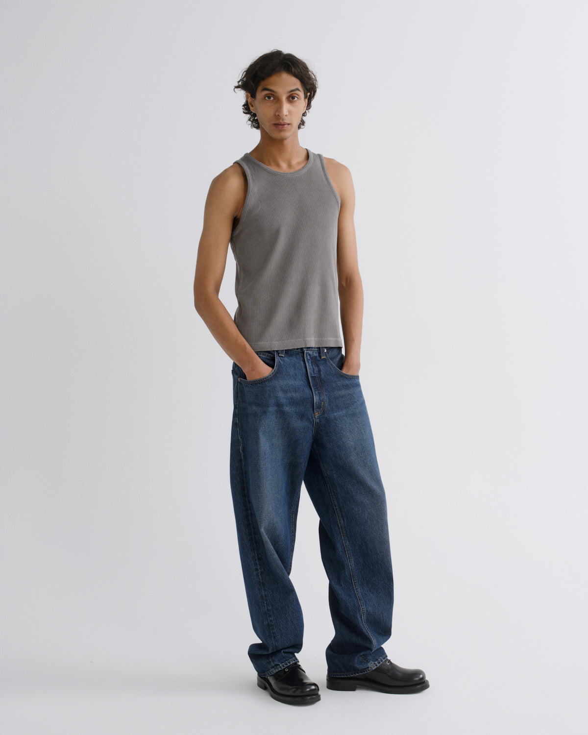 A model stands against a light gray studio background wearing a fitted gray sleeveless tank top, loose dark blue jeans, and black shoes.