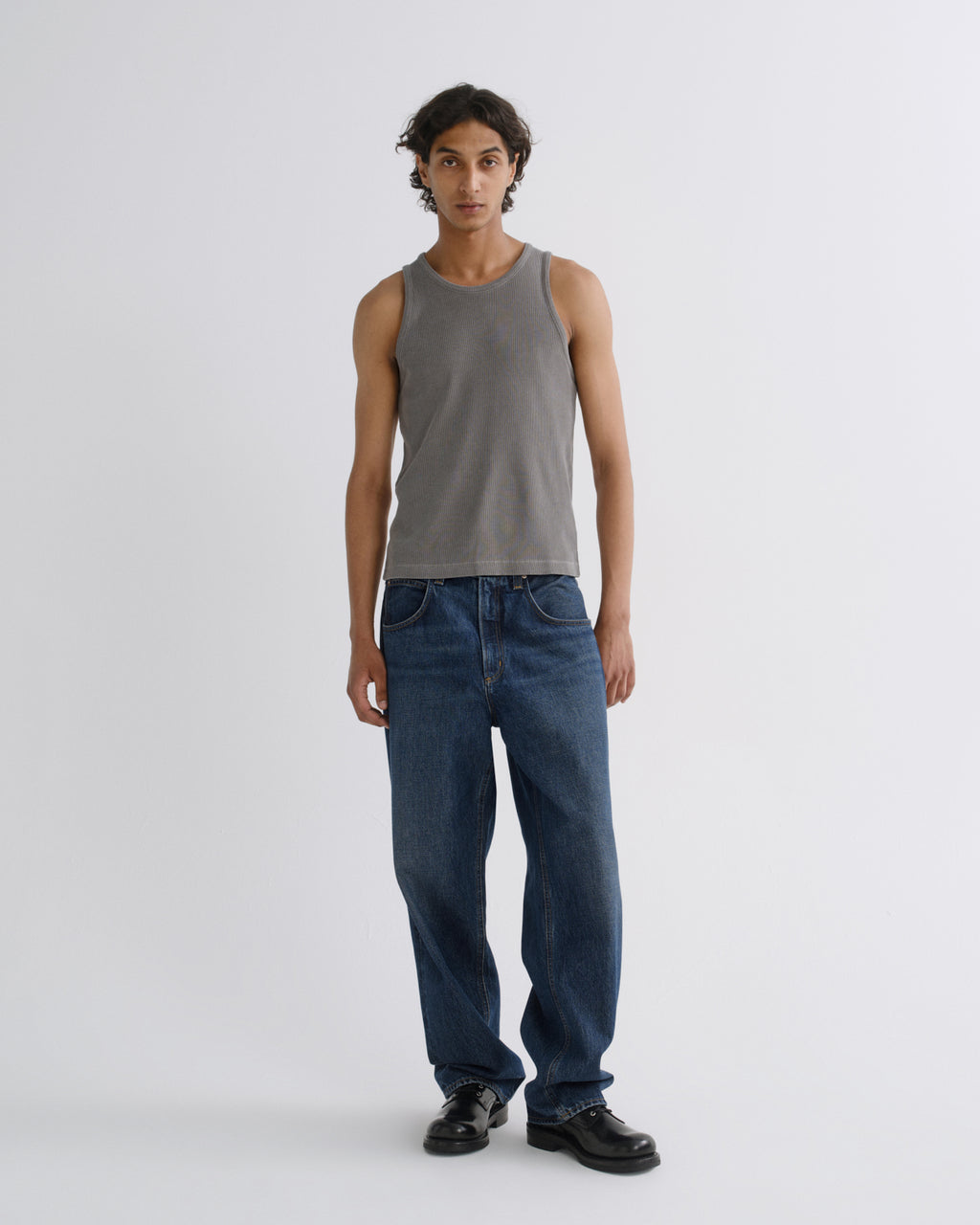 A model stands against a light gray studio background wearing a washed gray ribbed tank top, blue jeans, and black leather shoes.