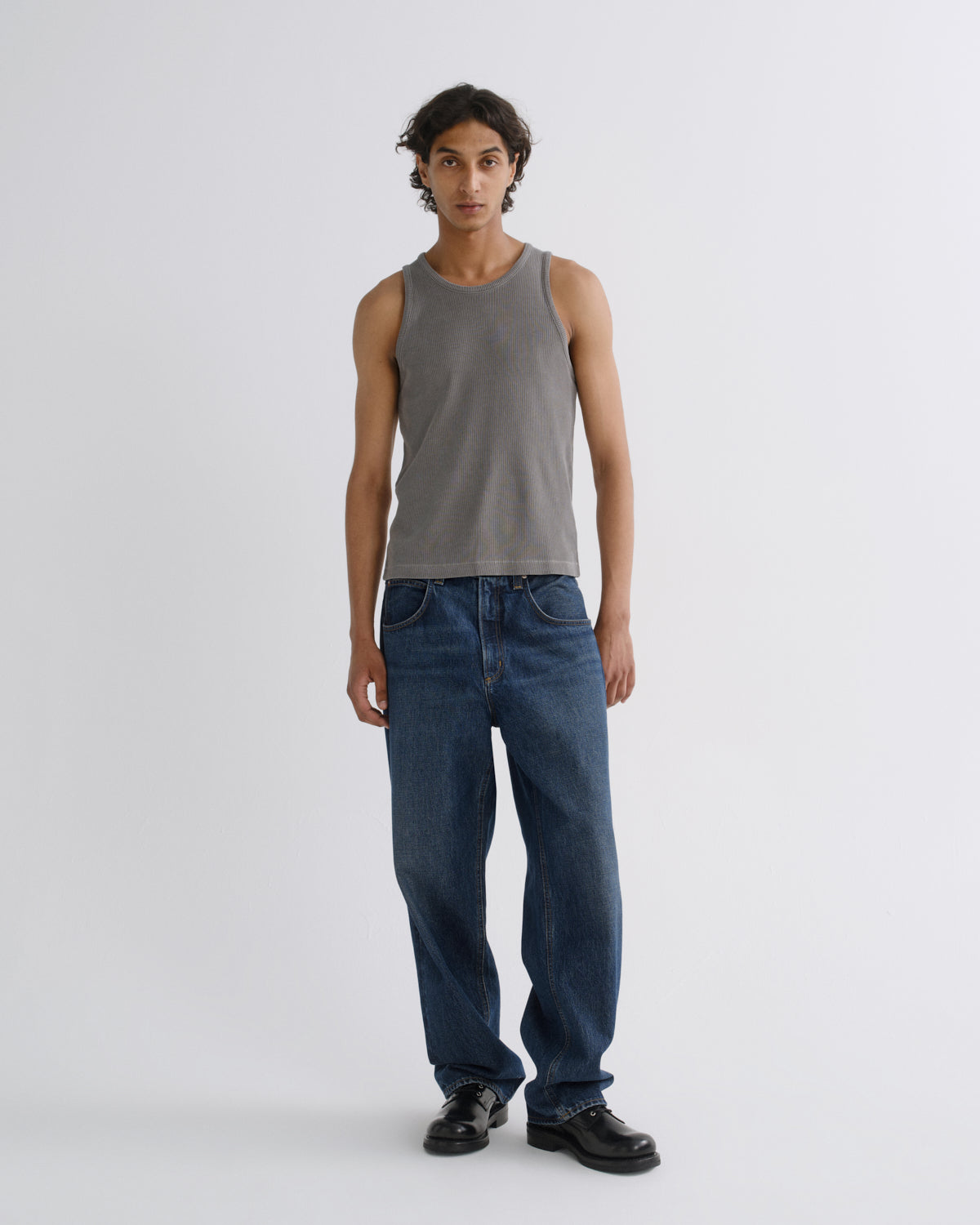 A model stands against a light gray studio background wearing a washed gray ribbed tank top, blue jeans, and black leather shoes.
