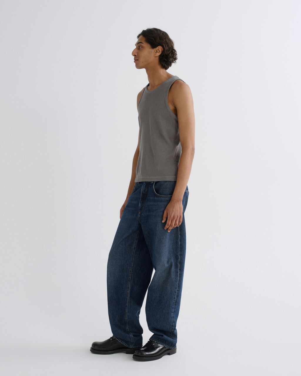 A man stands in profile wearing a gray sleeveless knit top, loose dark blue jeans, and black shoes against a plain light gray studio background.