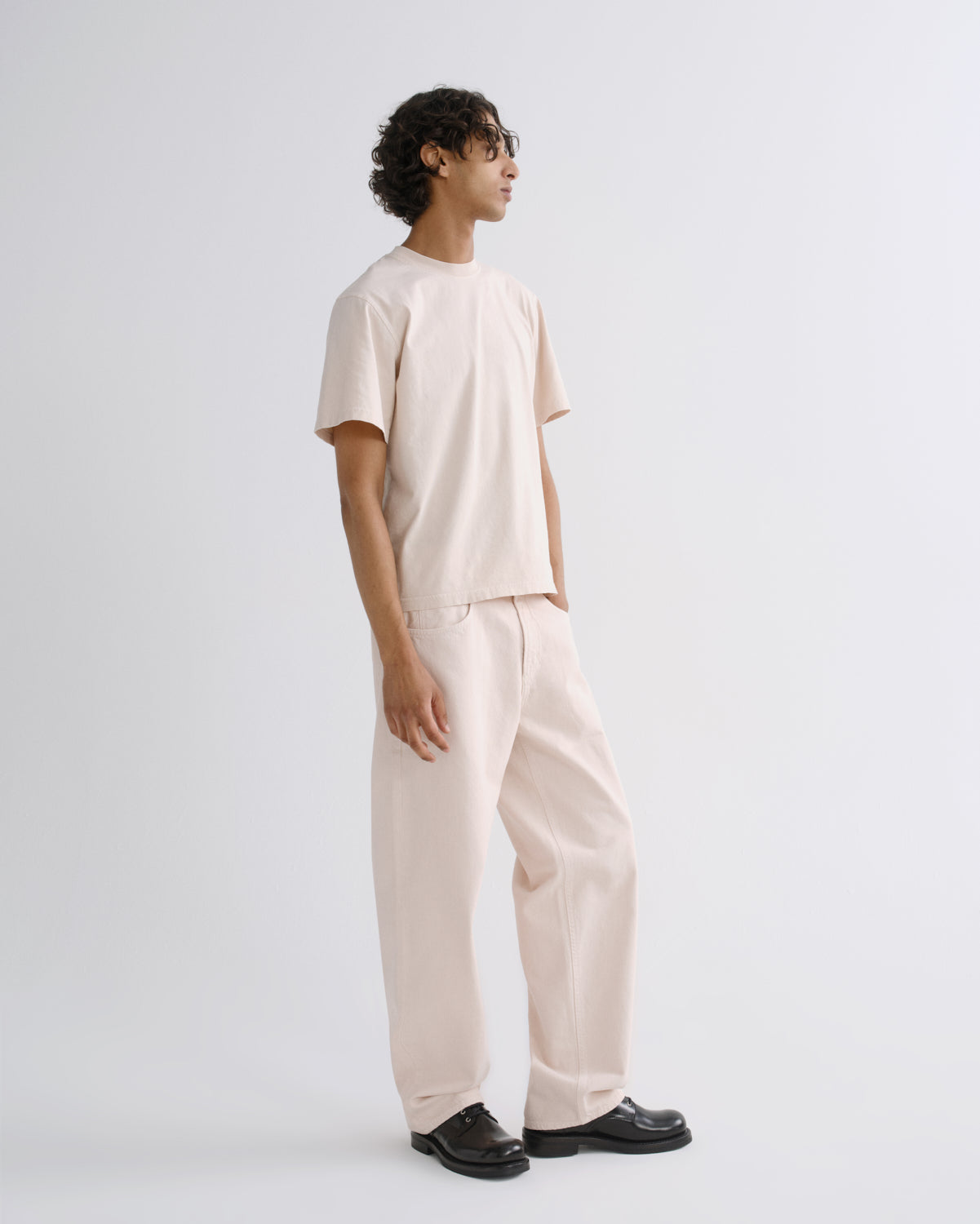 A model wearing a light beige short-sleeve t-shirt and matching wide-leg trousers, paired with black leather shoes, standing in profile against a plain light gray studio background.