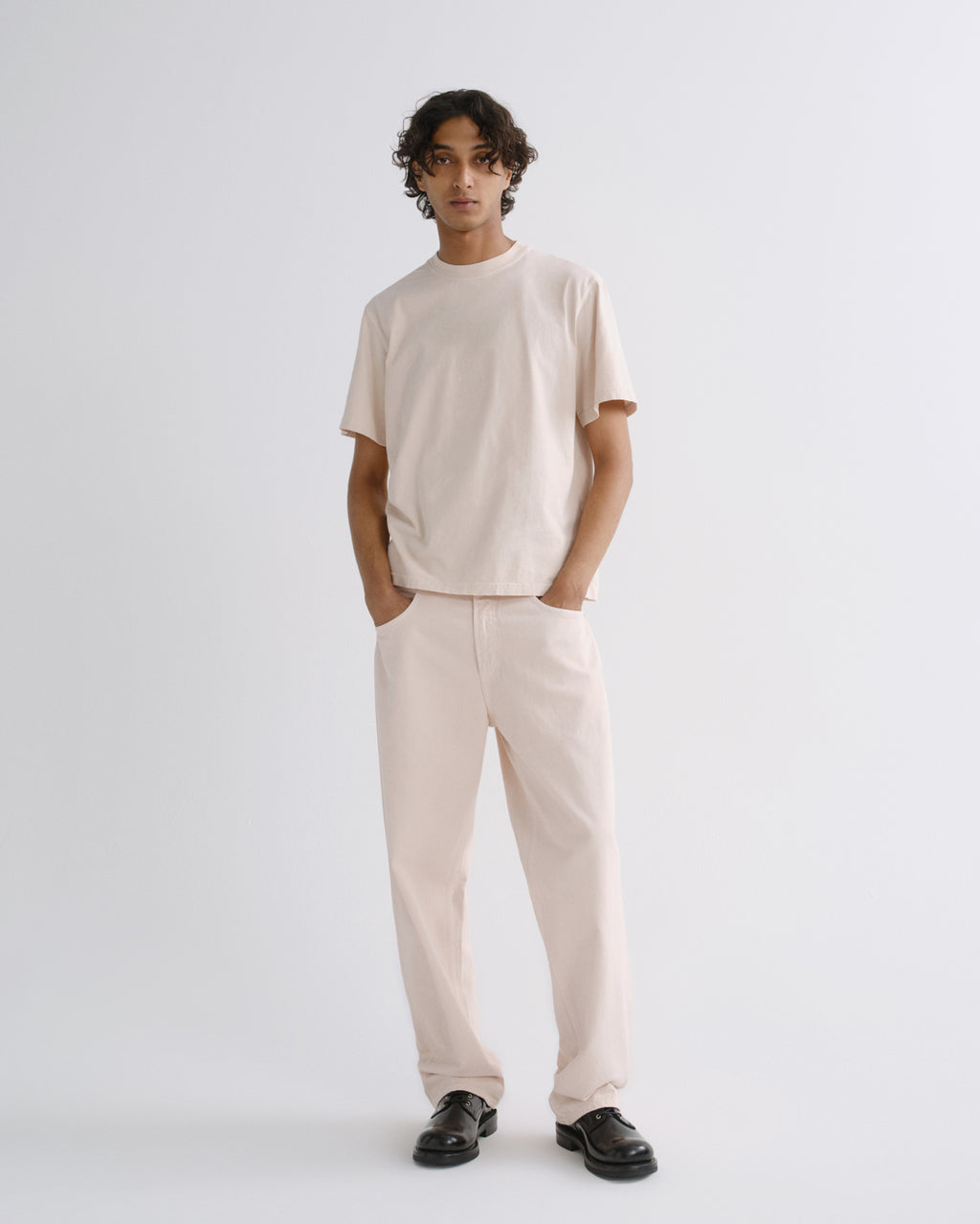 A model stands against a light gray studio background wearing a light beige short-sleeve crewneck t-shirt and matching light beige relaxed-fit trousers, paired with black loafers.