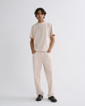 A model stands against a light gray studio background wearing a light beige short-sleeve crewneck t-shirt and matching light beige relaxed-fit trousers, paired with black loafers.
