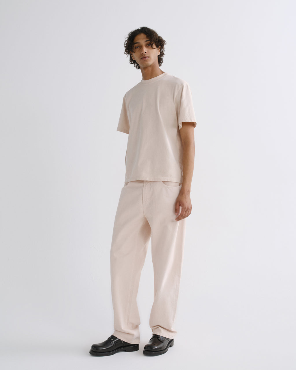 A model stands in a studio wearing a light beige short-sleeve t-shirt and matching wide-leg trousers, paired with black leather shoes, against a plain light gray background.