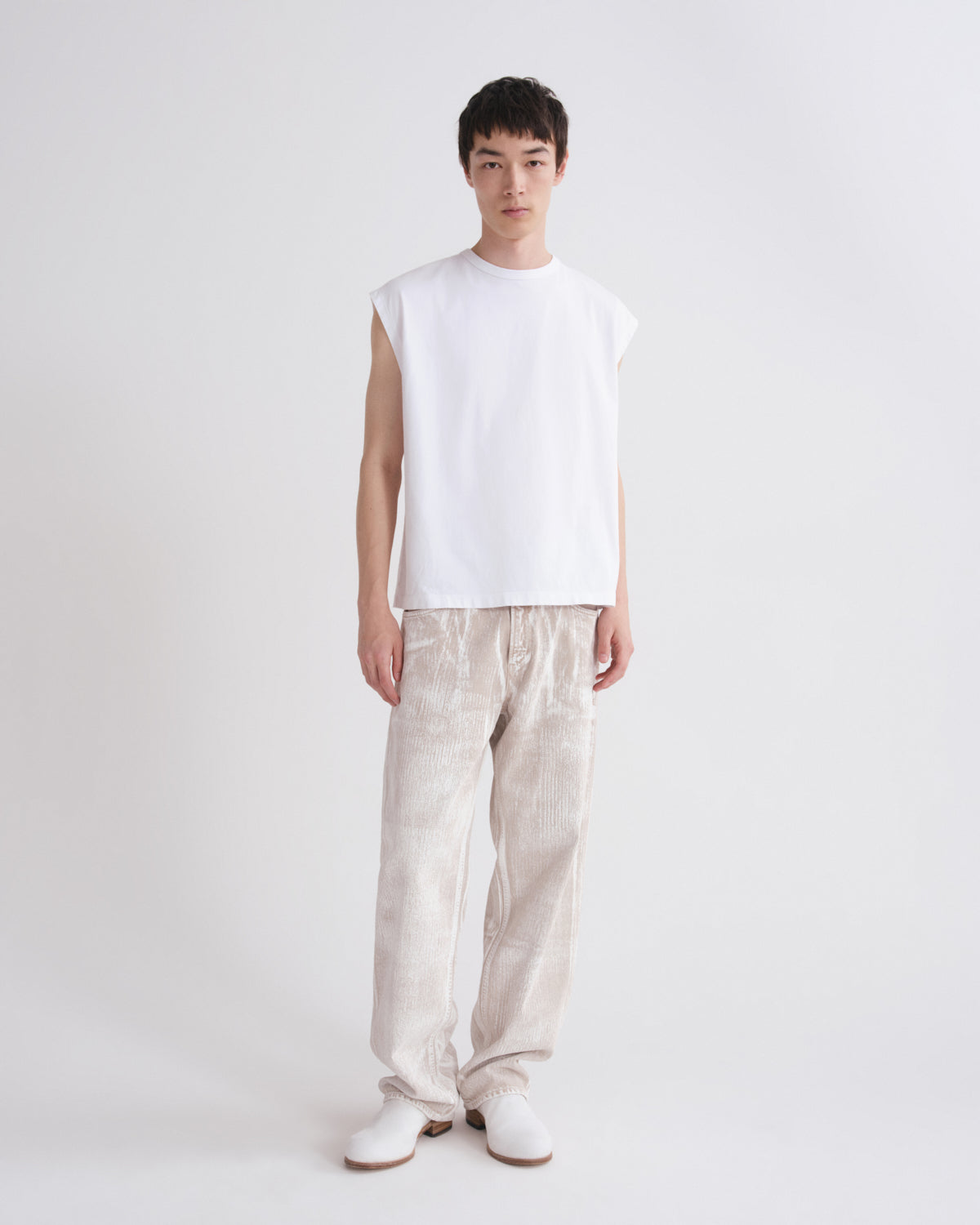 A model stands against a plain light gray studio background wearing a white sleeveless crewneck t-shirt, loose light beige trousers, and white shoes.