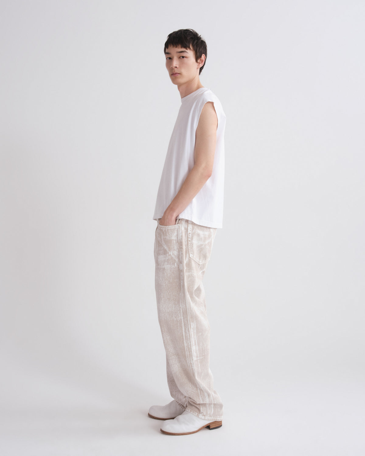 A model stands in profile wearing a white sleeveless top, light beige textured wide-leg trousers, and white shoes against a plain light gray studio background.