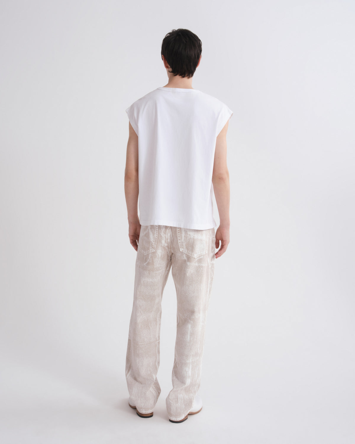 A model shown from the back wearing a white sleeveless t-shirt and light beige patterned trousers, standing against a plain white studio background.