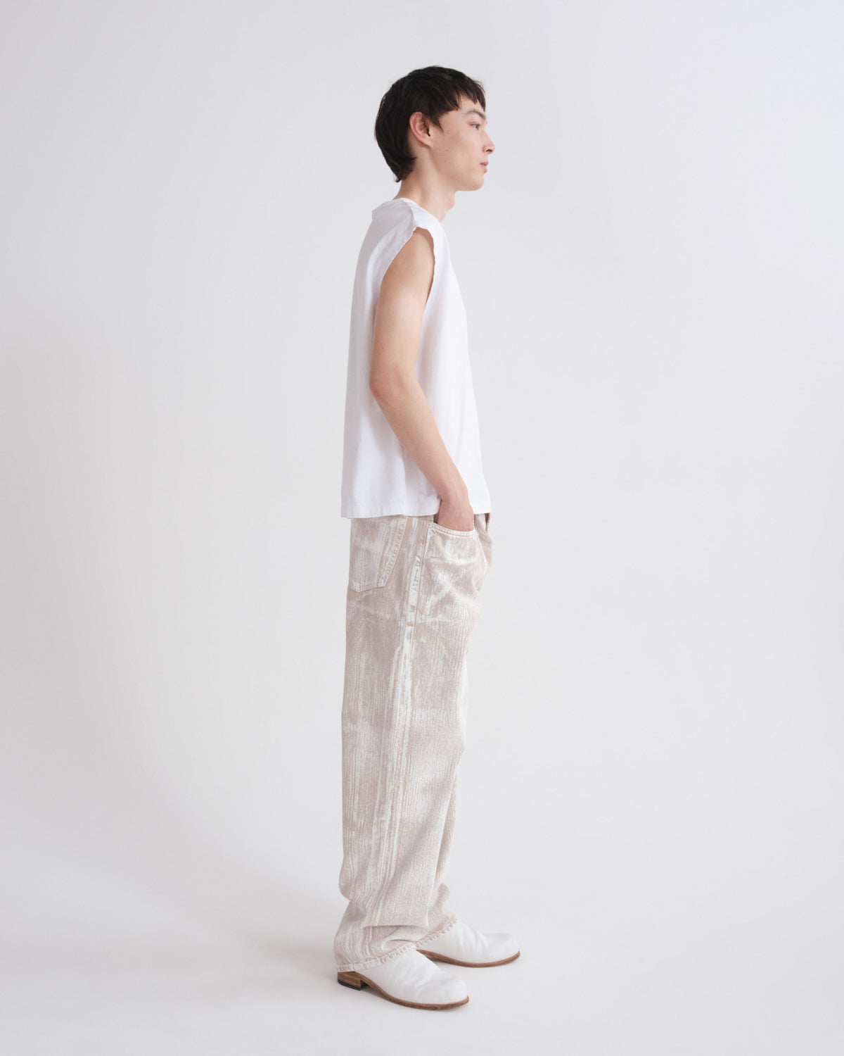 A model standing in profile wearing a sleeveless white top, cream textured pants, and white shoes against a plain light gray studio background.
