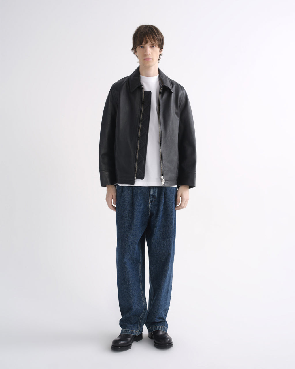 Person wearing a black leather jacket with blue jeans and black shoes on a grey background