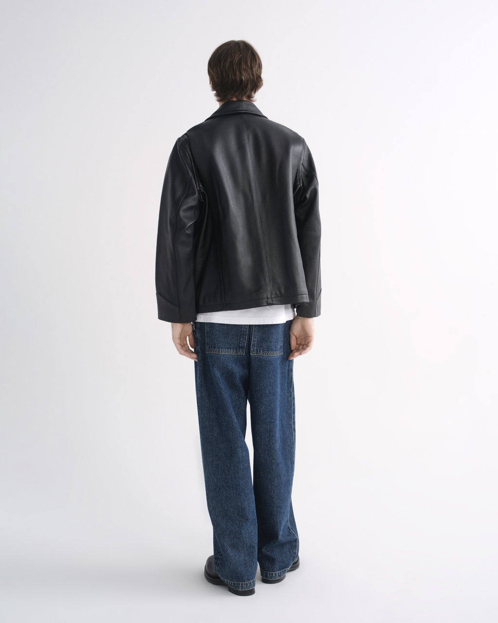 Person wearing a black leather jacket with blue jeans and black shoes, back view, on a grey background
