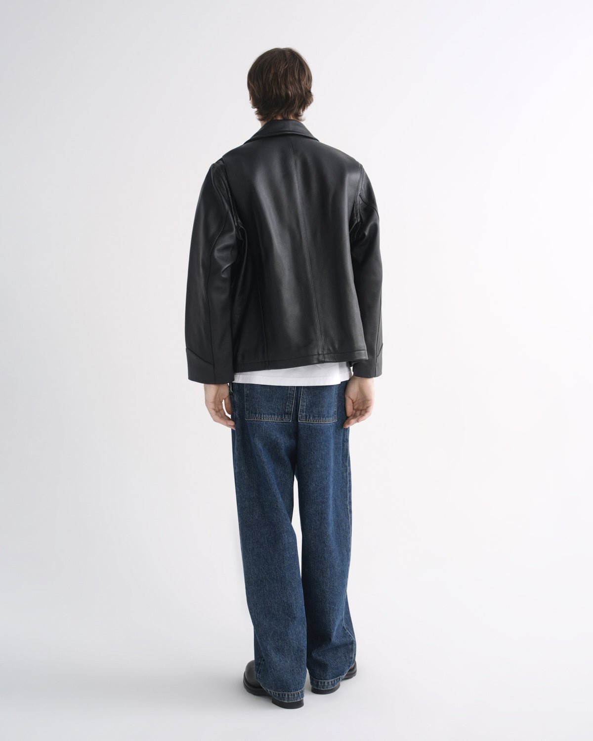 Person wearing a black leather jacket with blue jeans and black shoes, back view, on a grey background