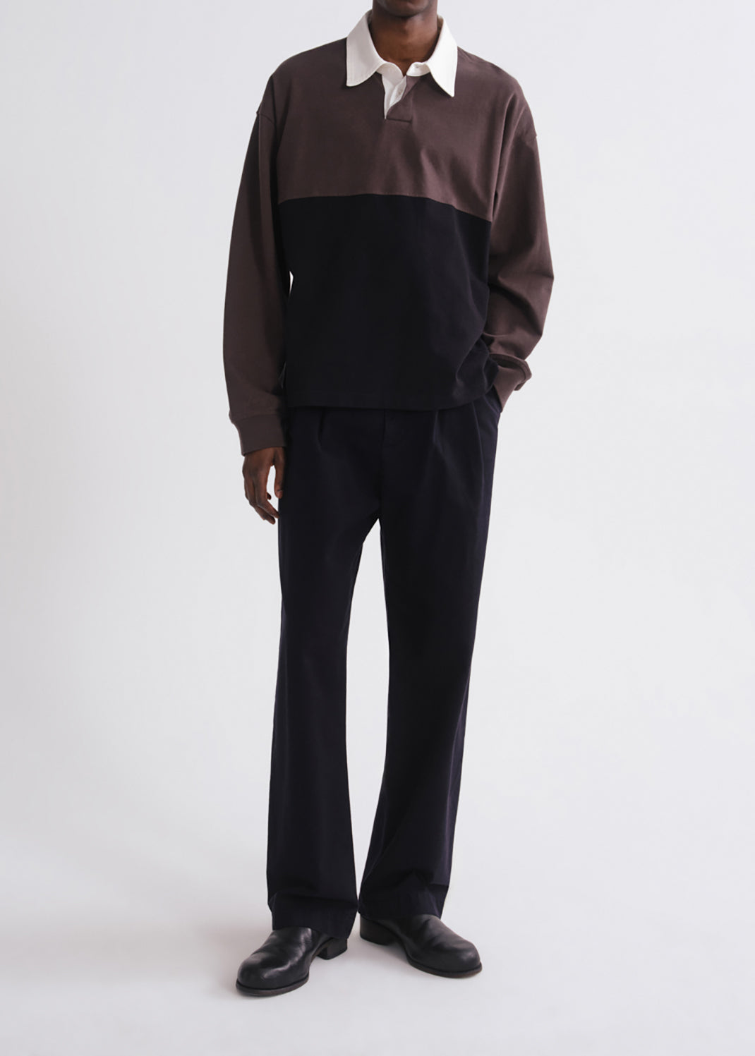 A model wearing a color-blocked long-sleeve rugby shirt with a brown and black body, a white collar, and black trousers, standing against a grey background.