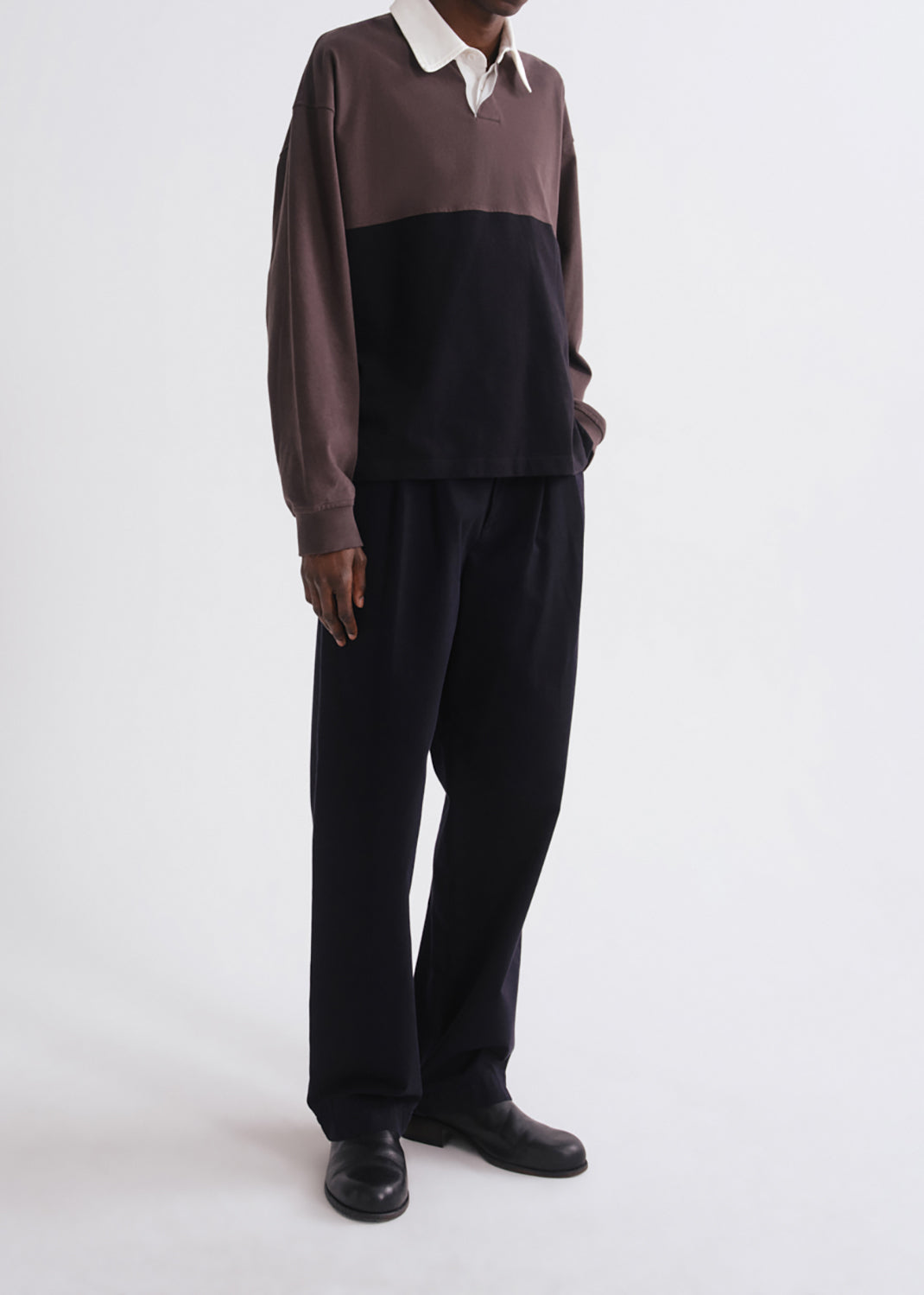 A model wearing a color-blocked long-sleeve rugby shirt in brown and black with a white collar, paired with black trousers and black shoes, shot against a grey background.