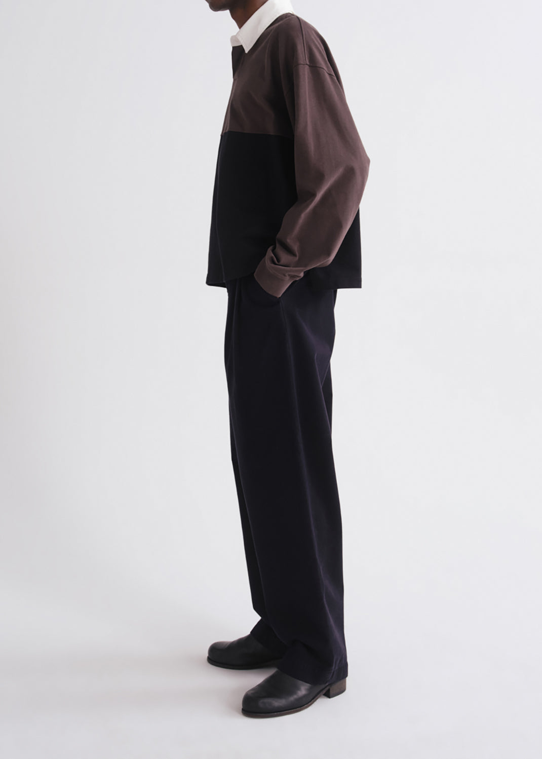 A model shown from the side wearing a color-blocked long-sleeve shirt with a brown upper section and dark lower section, paired with dark trousers and black shoes, against a grey background.