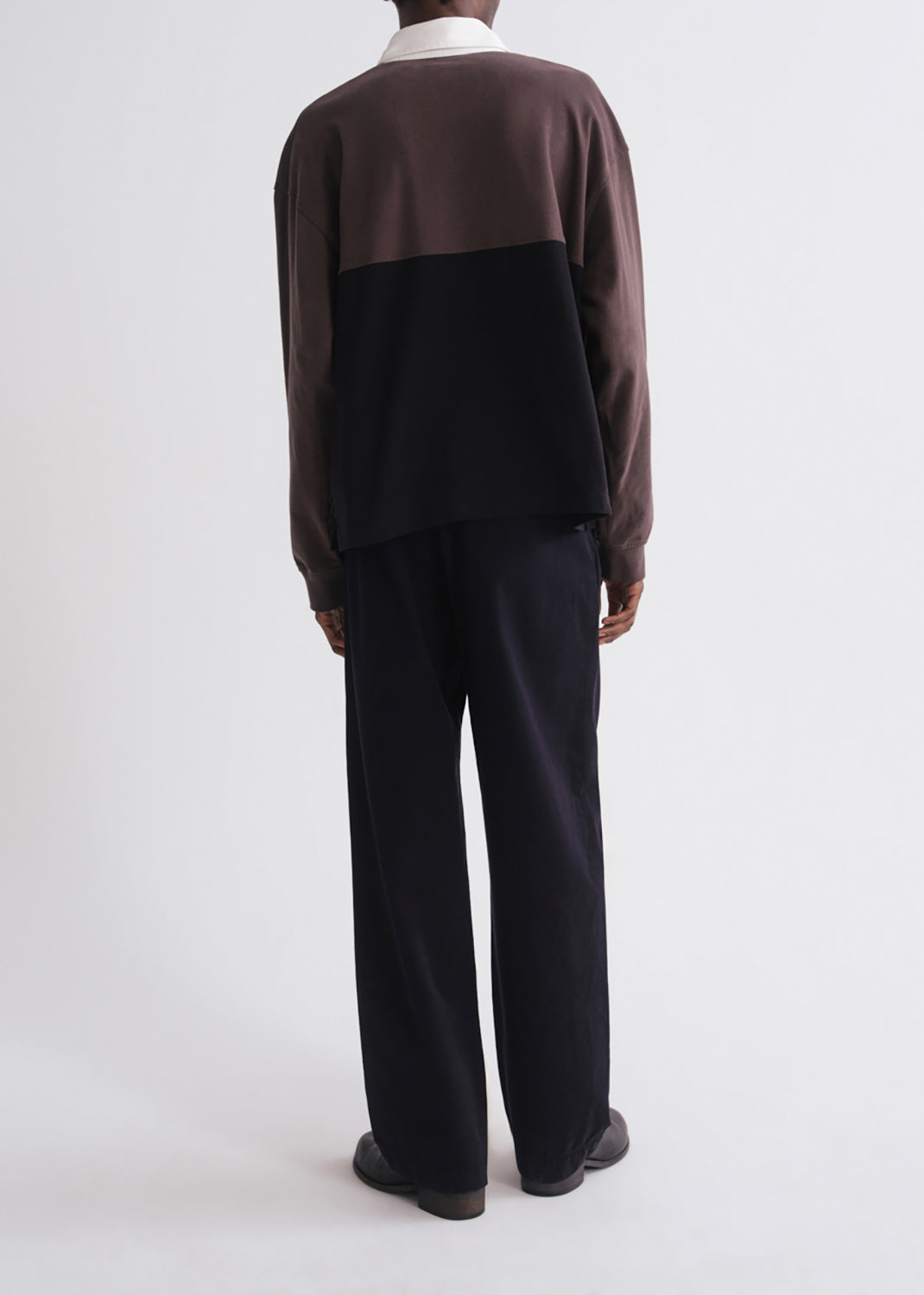A model viewed from the back wearing a color-blocked knit sweater with a brown upper section and sleeves, a black lower panel, and a white collar, paired with dark wide-leg trousers.