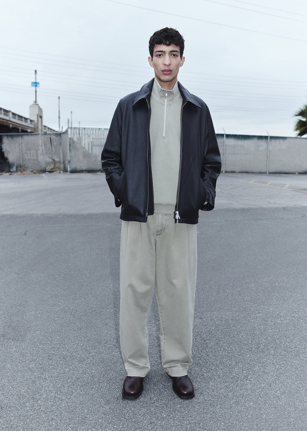 Man wearing a black jacket, beige shirt, and light-colored pants standing in an open area with a fence and sky in the background.