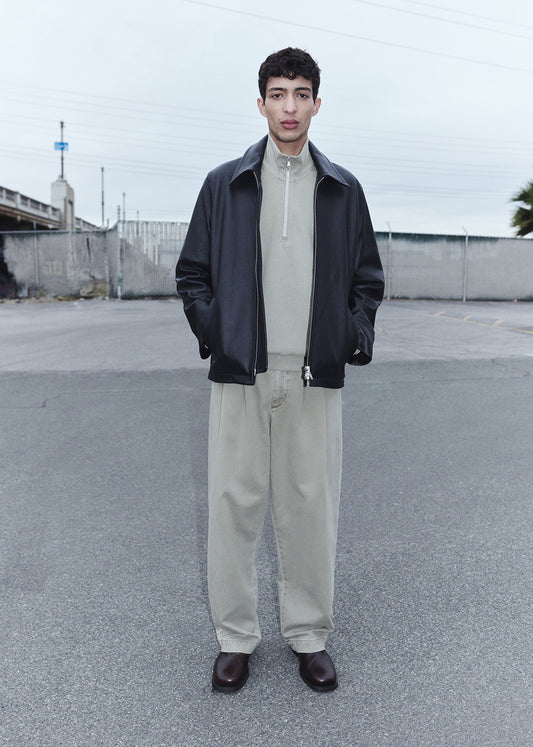 Man wearing a black jacket, beige shirt, and light-colored pants standing in an open area with a fence and sky in the background.