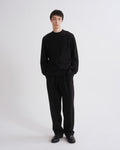 A man wearing a black long-sleeve crewneck sweater and matching black trousers, standing against a plain light gray studio background.