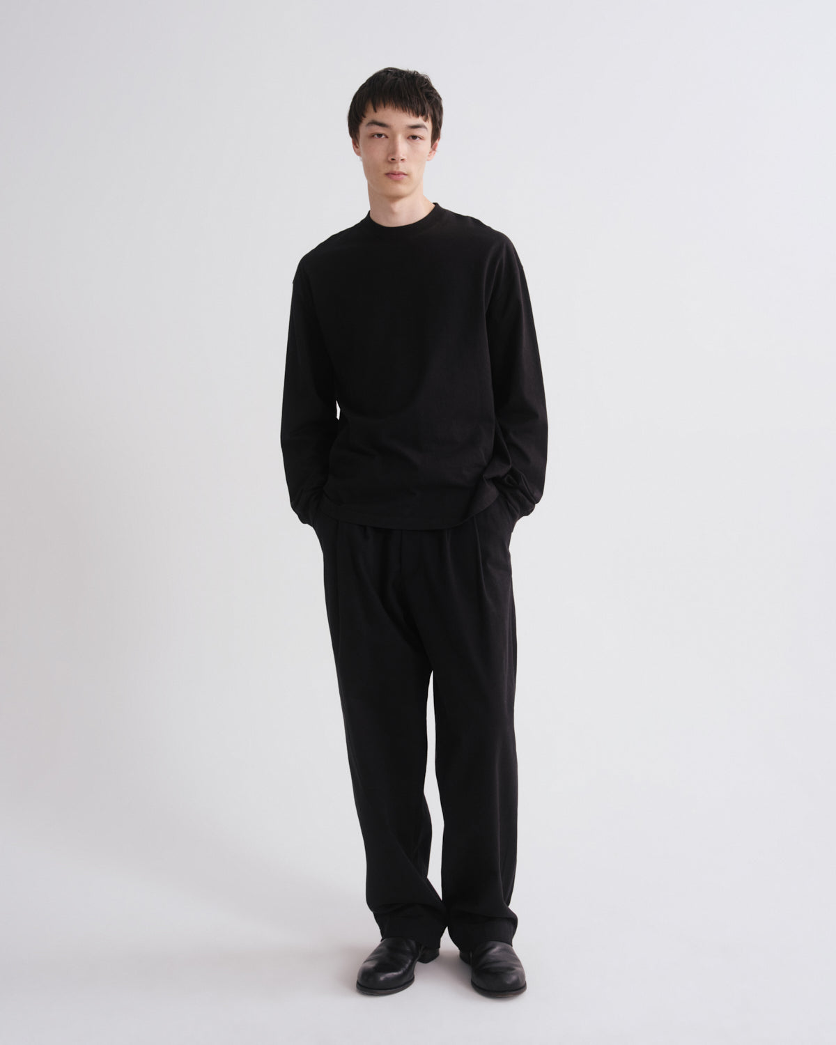 A man wearing a black long-sleeve crewneck sweater and matching black trousers, standing against a plain light gray studio background.